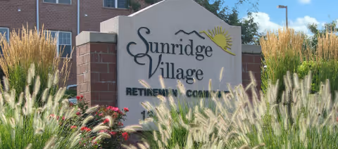 Stone sign for Sunridge Village Retirement Community surrounded by ornamental grasses and flowers with a brick building and blue sky in the background.