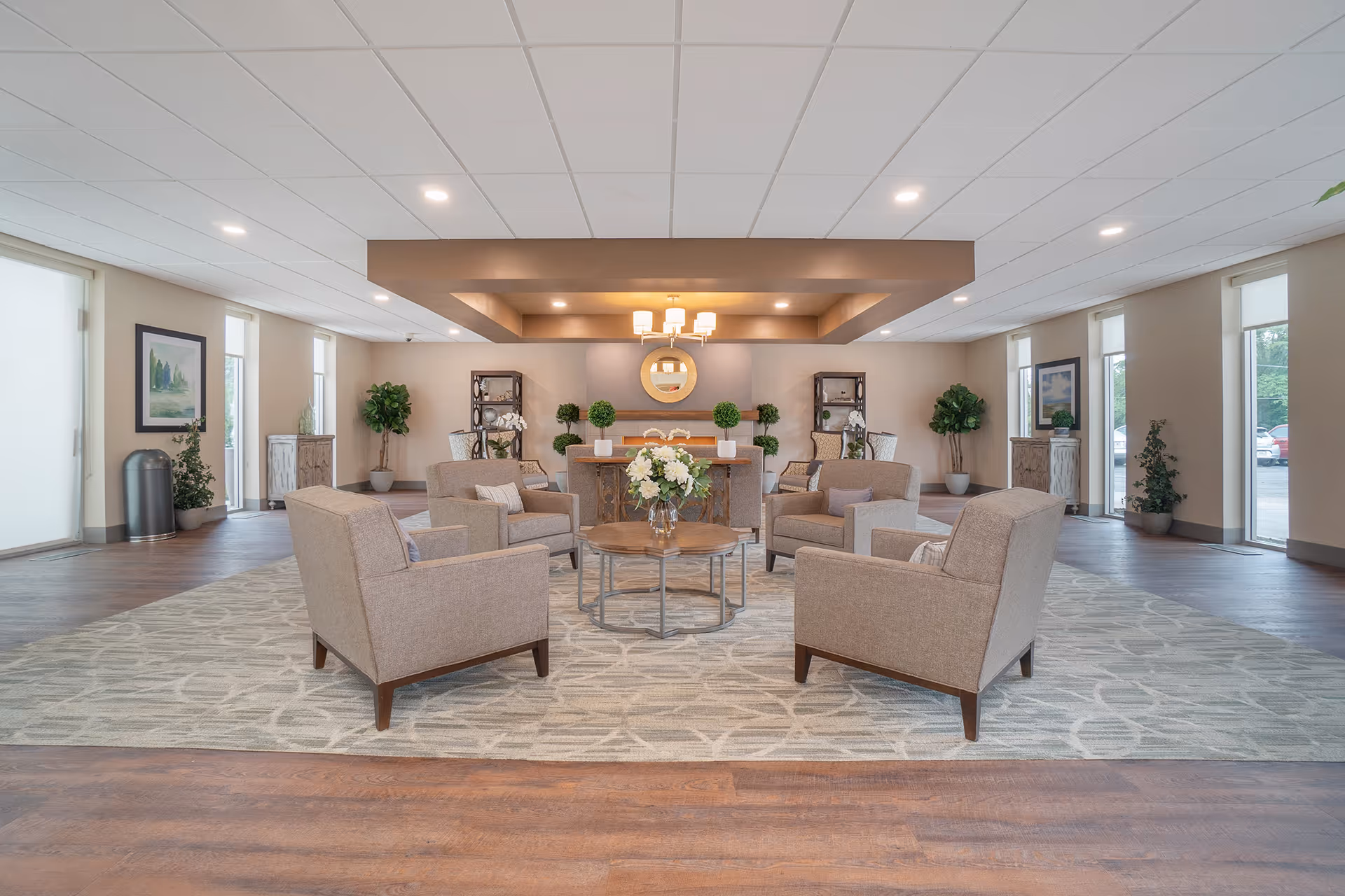 A spacious and well-lit common area with four beige armchairs arranged around a round wooden coffee table with a vase of white flowers. The room features a large patterned rug, wooden flooring, potted plants, framed artwork on the walls, and a decorative ceiling with recessed lighting and a chandelier.