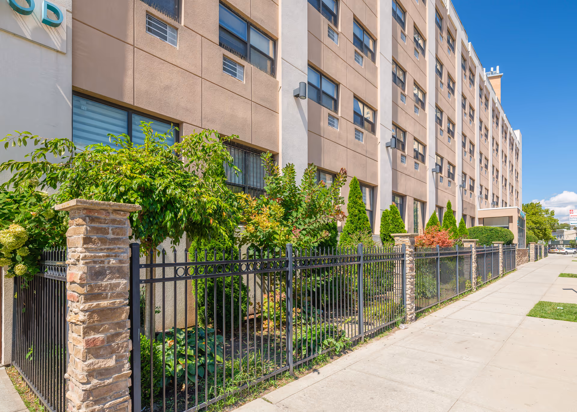 Exterior view of a multi-story building with beige and white walls, several windows, and a black metal fence with stone pillars in front. There are green shrubs and small trees planted along the fence, and a clear blue sky overhead.