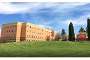 Exterior view of a large, multi-story brick building under a blue sky with some clouds. The building is surrounded by a well-maintained green lawn and a few tall trees. There is a sign on the right side of the image indicating the facility name.