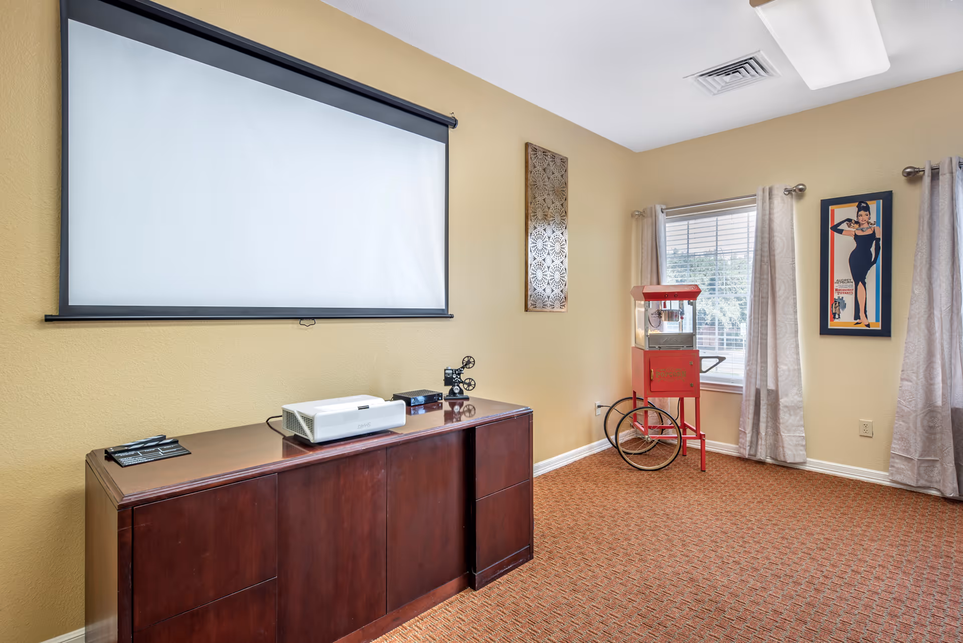 A room with a large white projection screen mounted on a beige wall above a dark wooden cabinet. On the cabinet, there is a white projector and a small decorative film camera. To the right, there is a vintage red popcorn machine on wheels near a window with beige curtains. A framed poster of a woman in a black dress is hung on the wall next to the window. The floor is carpeted in a patterned brown color.