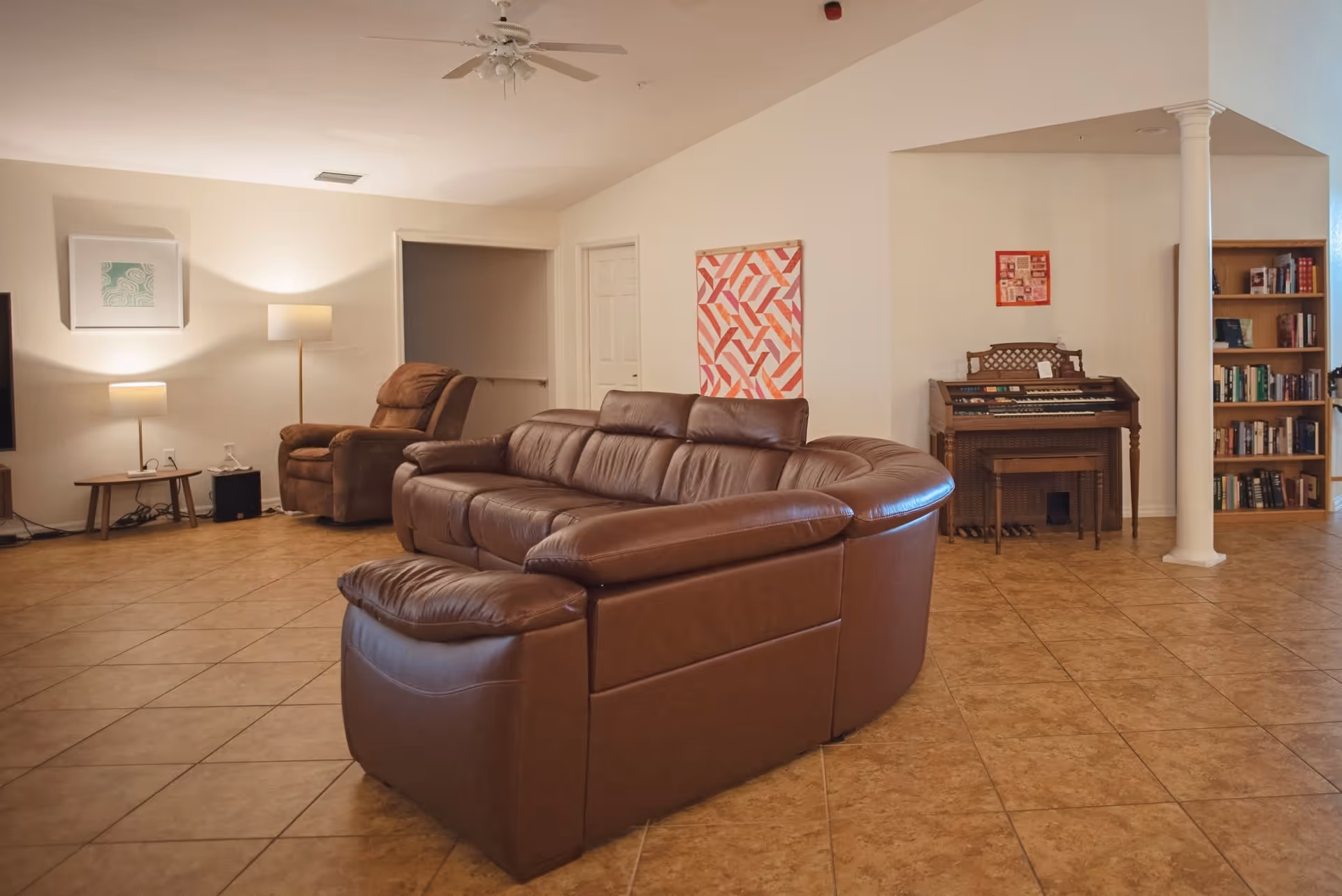 A spacious living room with a large brown leather sectional sofa in the center. To the left, there is a brown recliner chair next to a small table with two lamps. On the right side, there is an organ with a bench and a bookshelf filled with books. The floor is tiled, and the walls are painted white with some colorful artwork hanging. A white ceiling fan is visible above.