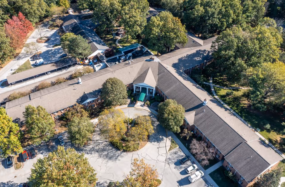 Aerial view of The Grove at Oakleaf Village of Sylvania, showing a large building with a circular driveway entrance surrounded by trees and greenery. Several cars are parked near the building, and the area is lush with mature trees and landscaped grounds.
