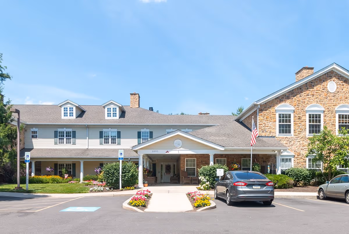 Front exterior view of Park Creek Place, a senior living facility with a combination of stone and siding facade, a covered entrance with columns, landscaped flower beds, and a parking area with cars.
