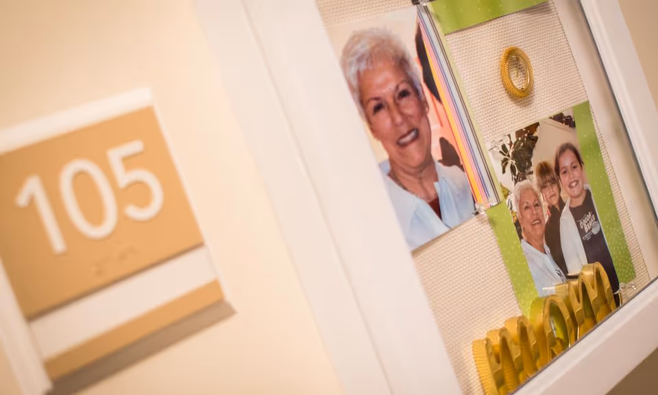 Close-up view of a senior living facility room door labeled 105 with a display case beside it containing photographs of an elderly woman and children, along with decorative items including the word 'MAMA' in gold letters.