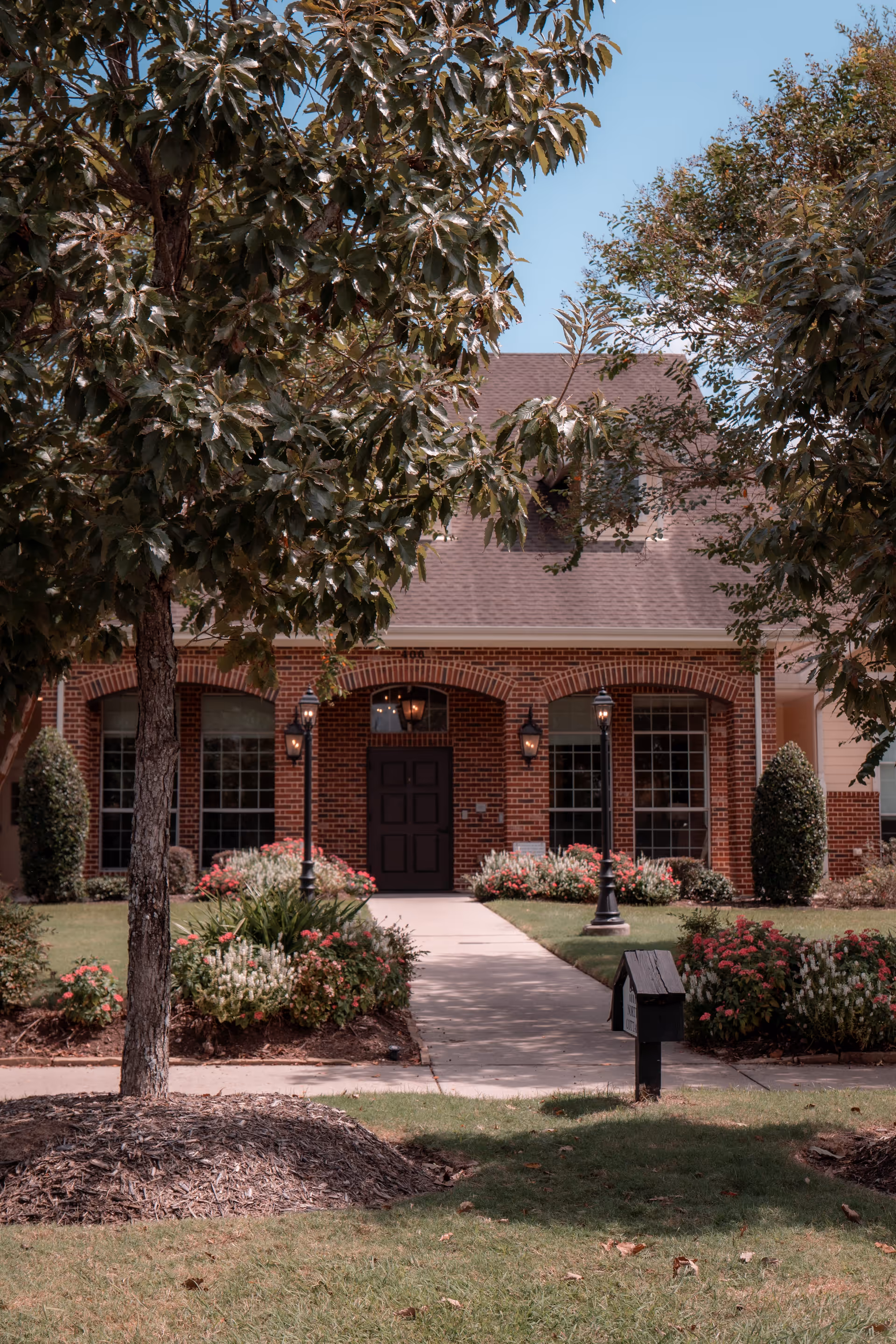 Front exterior view of a brick building with a dark door, large windows, and a sloped roof. The entrance is framed by three arches and flanked by two black lamp posts. The walkway leading to the door is bordered by well-maintained flower beds with pink and white flowers, green shrubs, and trees providing partial shade.