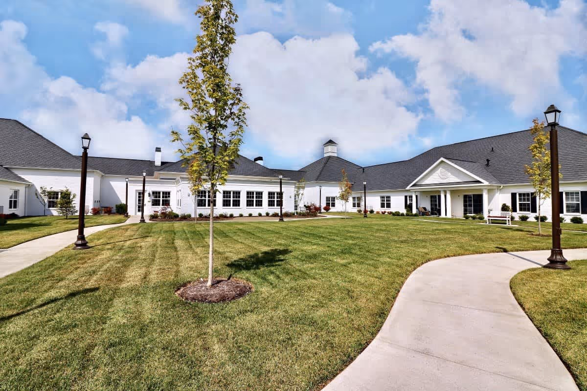 Exterior view of Cherry Blossom Senior Living facility showing a large, well-maintained lawn with young trees and a curved concrete walkway leading to a white building with a dark roof under a partly cloudy sky.