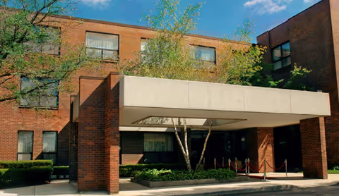 Exterior view of a brick building with multiple windows, featuring a covered entrance supported by brick pillars and a small landscaped area with trees and shrubs under a clear blue sky.