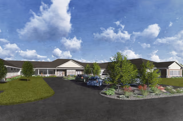 Exterior view of a single-story senior living facility building with a driveway leading to the entrance. Several cars are parked near the entrance, and there are landscaped areas with trees and shrubs around the driveway. The sky is partly cloudy with blue patches.