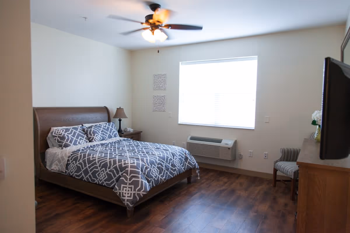 A bedroom in a senior living facility with a wooden bed frame, patterned gray and white bedding, a nightstand with a lamp and phone, a window with blinds, a wall-mounted air conditioning unit, a striped armchair, a wooden dresser with a TV on top, and a ceiling fan with lights.