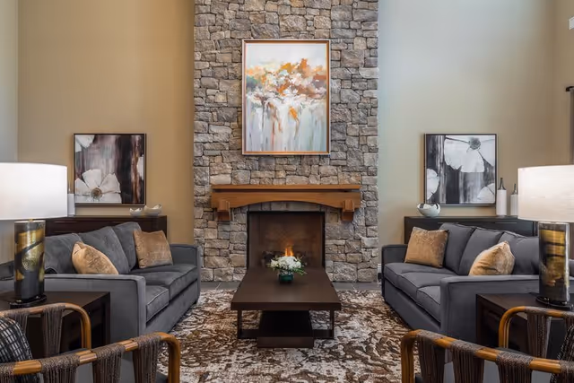 A cozy living room with two gray sofas facing each other, each with beige and brown throw pillows. Between the sofas is a dark wooden coffee table with a small flower arrangement. The room features a stone fireplace with a wooden mantle and a colorful abstract painting above it. On either side of the sofas are dark wooden side tables with modern lamps. The walls are painted in neutral tones, and there are two black and white floral paintings on the walls behind the sofas. The floor is covered with a patterned area rug.