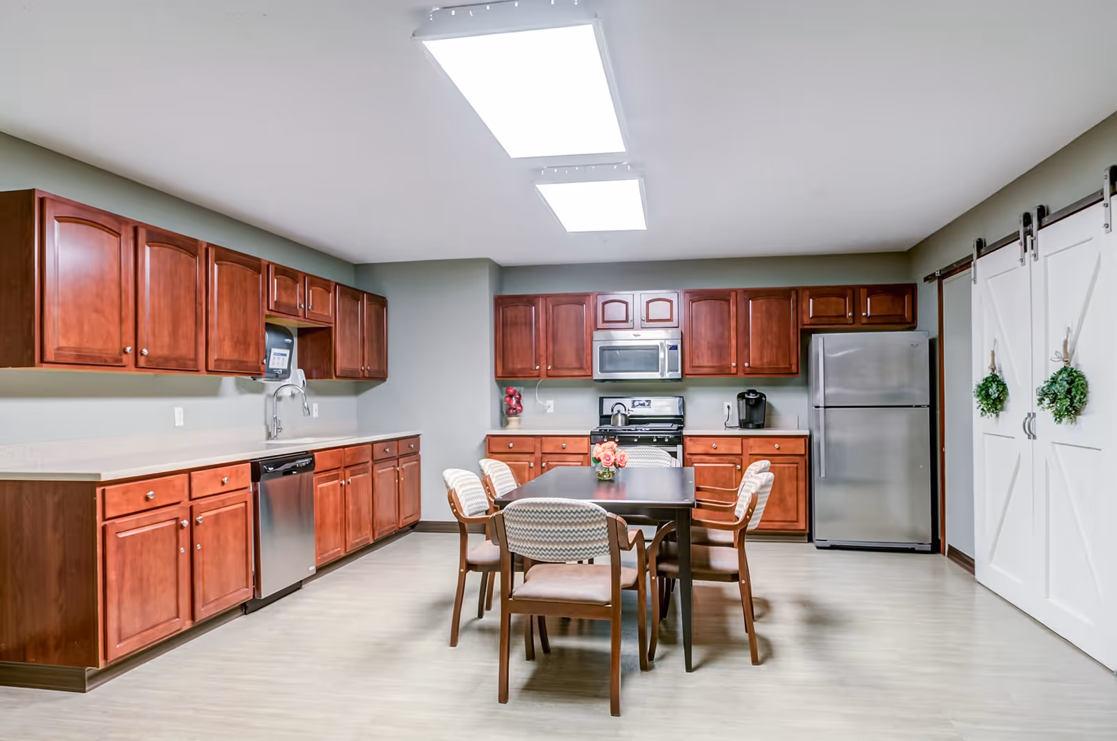 A spacious kitchen with wooden cabinets, stainless steel appliances including a refrigerator, dishwasher, microwave, and stove. There is a rectangular dining table in the center with six cushioned chairs around it. The room has bright overhead lighting and two white sliding barn doors decorated with small green wreaths.