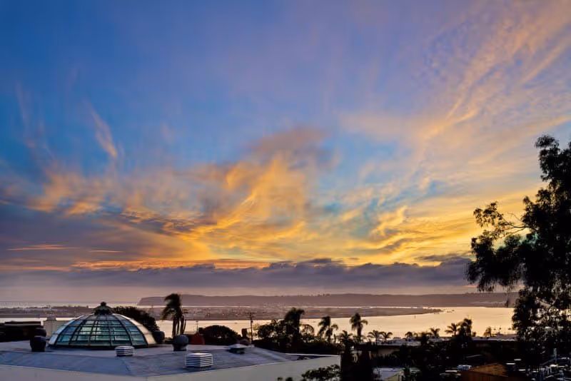 A scenic view at sunset showing a colorful sky with orange, yellow, and blue hues above a body of water. In the foreground, there is a rooftop with a glass dome structure and some palm trees, with more trees and buildings visible in the distance.
