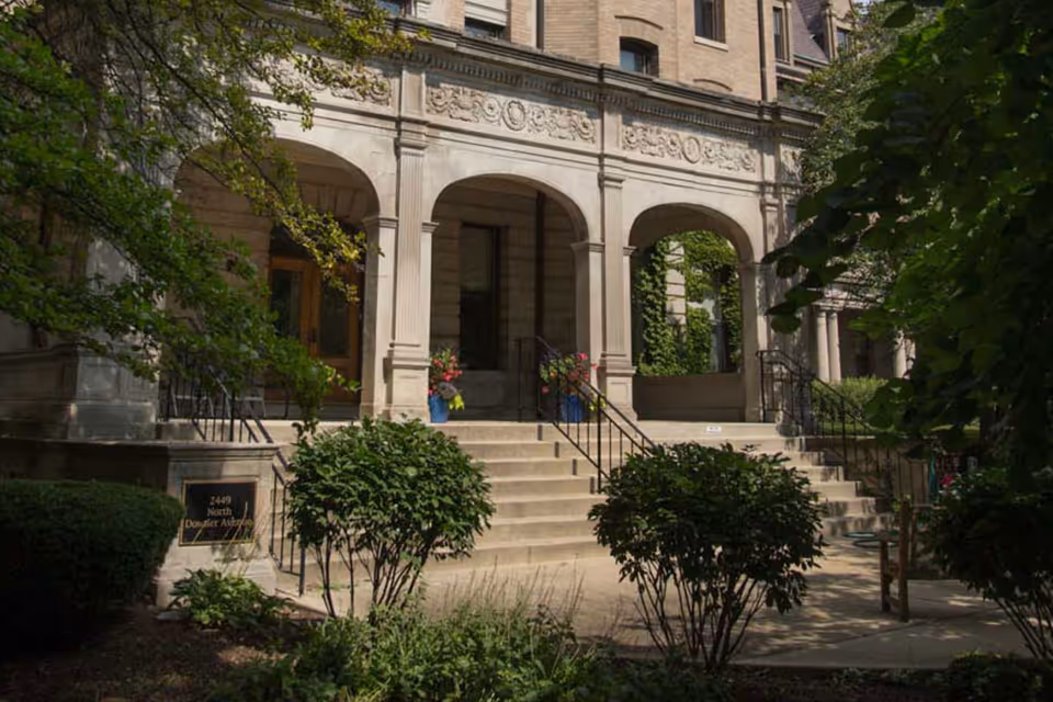 Front exterior view of a classic stone building with three arched entrances, stairs leading up to the doors, and greenery including bushes and trees surrounding the entrance area.