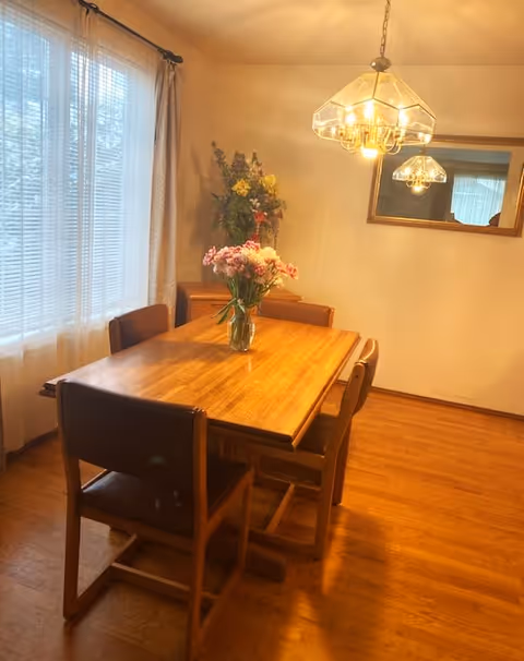 A dining room with a wooden table and four chairs. A vase with pink and white flowers is placed on the table. There is a window with sheer curtains on the left side, a wooden floor, a hanging light fixture above the table, and a mirror on the wall reflecting the light fixture. A small cabinet with a floral arrangement is in the corner.