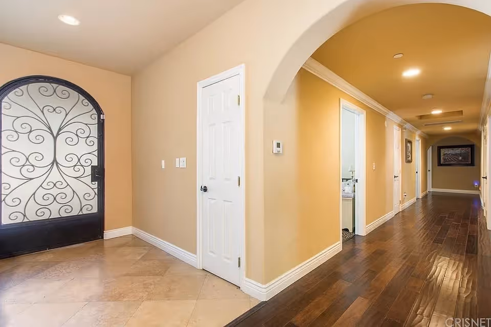 Interior view of a hallway in a senior living facility with beige walls, a decorative wrought iron door on the left, a white door in the center, and a long corridor with wooden flooring and recessed ceiling lights.