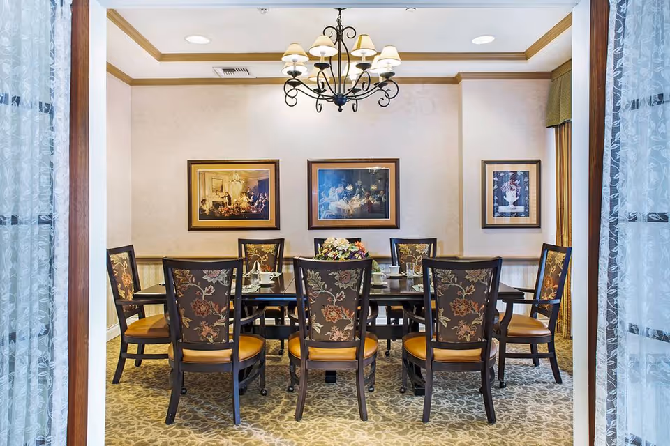 A formal dining room with a rectangular dark wood table surrounded by eight chairs with floral upholstery and tan seats. A chandelier with six lamps hangs above the table. Three framed paintings are displayed on the beige walls. The room has patterned carpet flooring and is viewed through a doorway with sheer white curtains on either side.