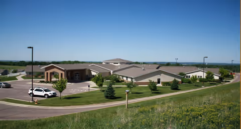 Exterior view of a single-story senior living facility building with a parking lot, several small trees, and a grassy area under a clear blue sky.