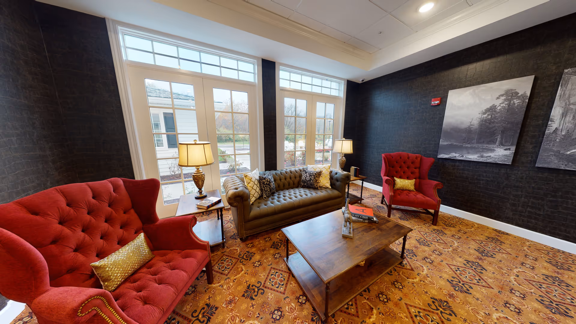 A cozy living room with two red tufted armchairs and a dark green tufted sofa arranged around a wooden coffee table. The room has large windows with white frames letting in natural light, dark textured walls, a patterned carpet, two table lamps on side tables, and black and white landscape artwork on the walls.