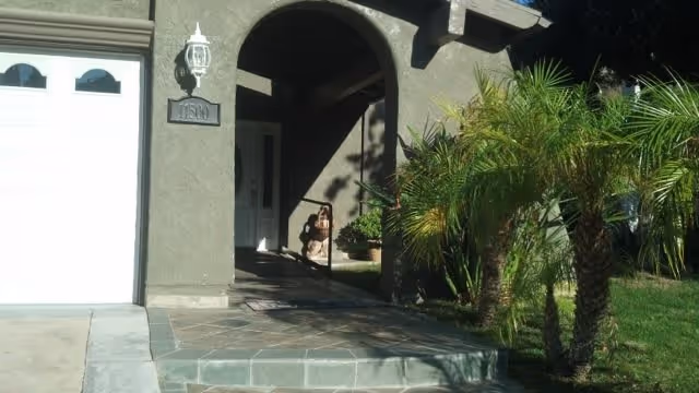 Front entrance of a residential building with an arched doorway, a white garage door on the left, a small porch area with a potted plant, and palm trees on the right side of the walkway.