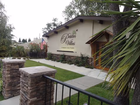 Exterior view of Palm Villas Memory Care Campbell facility showing the building facade with the facility name and logo, a walkway leading to the entrance, a small lawn area, stone pillars, and an American flag.