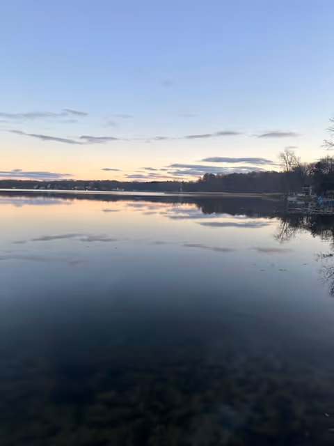 Calm lake at sunset with a clear sky and scattered clouds, reflecting on the still water. Trees line the far shore and the right side of the lake, with some houses or docks visible among the trees.