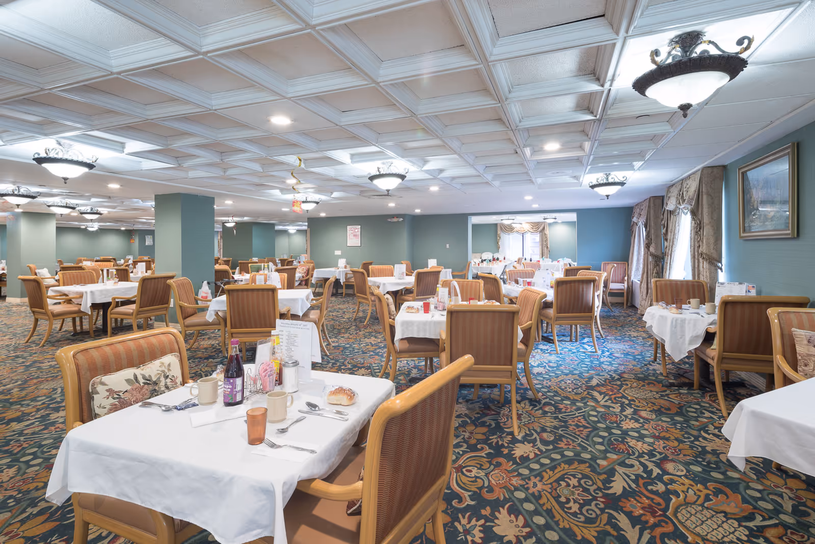 A spacious dining room with multiple tables covered in white tablecloths, each set with cups, plates, and utensils. The room features patterned carpet, green walls, large windows with drapes, and decorative ceiling lights.