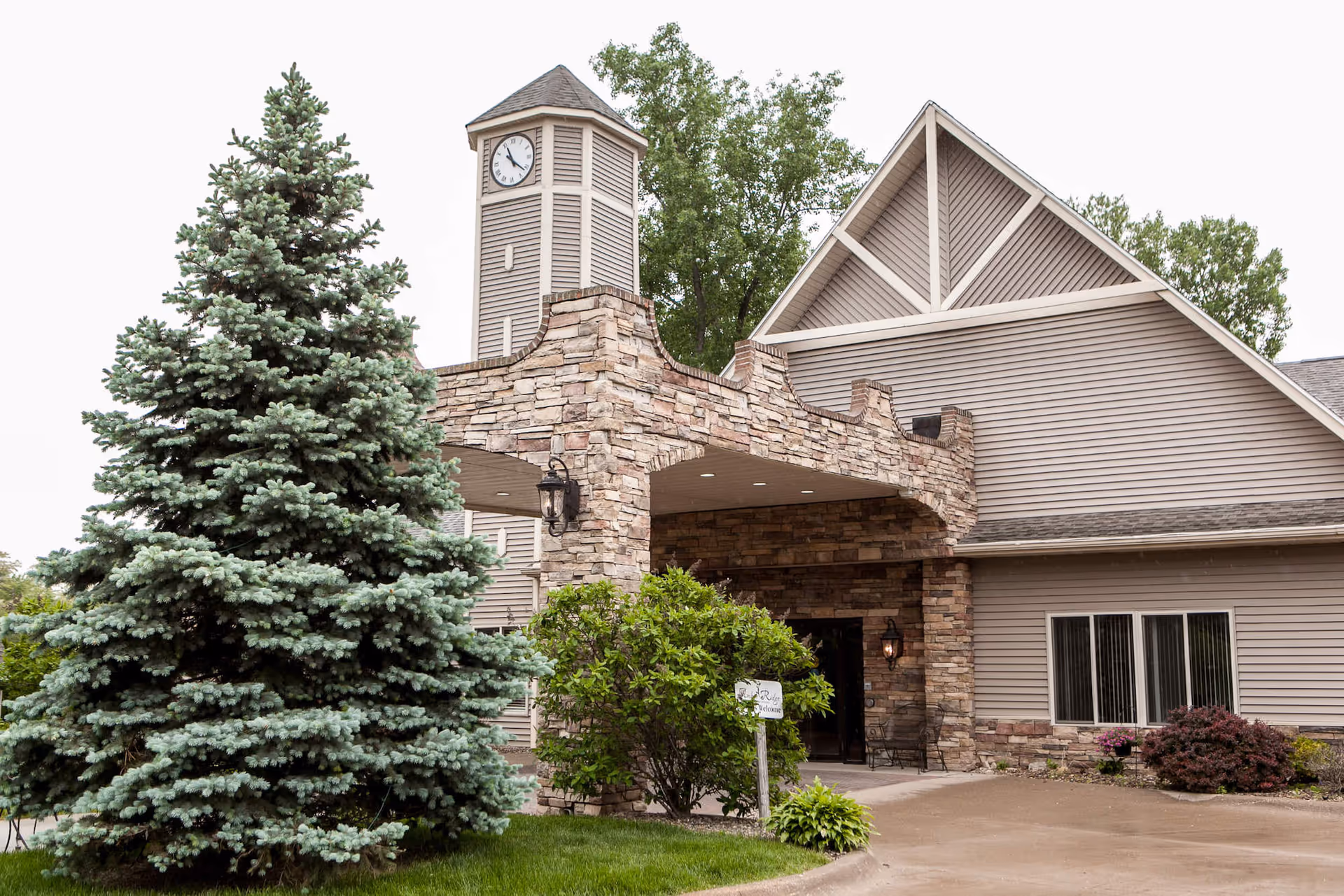 Exterior view of Charter Senior Living of Moline featuring a stone and siding building with a clock tower, surrounded by trees and shrubs, with a covered entrance area.