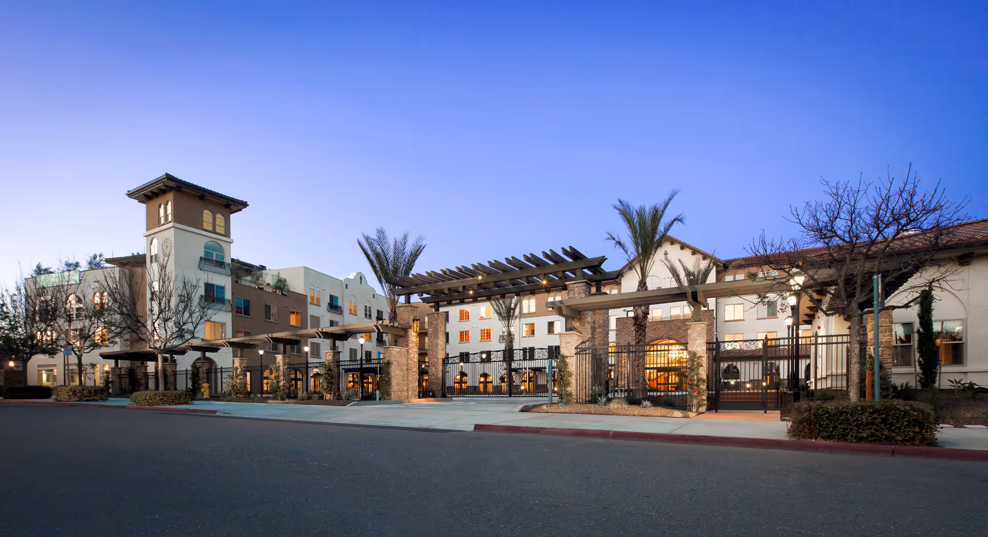 Exterior view of St. Paul's Plaza senior living facility at dusk, featuring a multi-story building with a clock tower, palm trees, stone pillars, and a gated entrance under a clear blue sky.