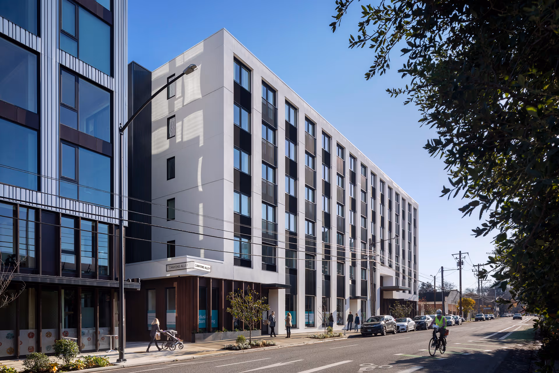 Street view of a modern multi-story building with large windows and people walking on the sidewalk. A cyclist is riding on the street, and there are parked cars along the road. Trees partially frame the right side of the image under a clear blue sky.