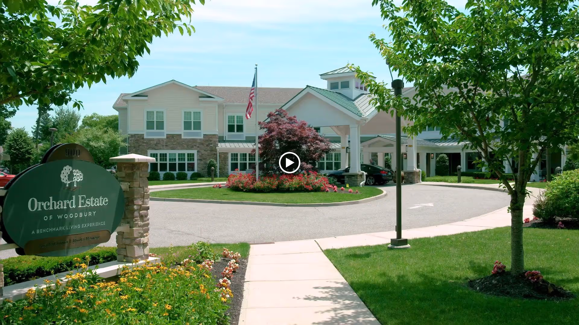 Front exterior view of Orchard Estate of Woodbury, a senior living facility, showing a two-story building with a covered entrance, landscaped garden with flowers and trees, an American flag, and a sign with the facility's name.
