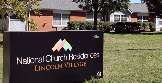 Outdoor view of a single-story brick building with white-framed windows and a gray roof. In the foreground, there is a black sign with colorful geometric shapes and the text 'National Church Residences Lincoln Village' along with the number 4959. The area around the building is grassy with some bushes and trees.