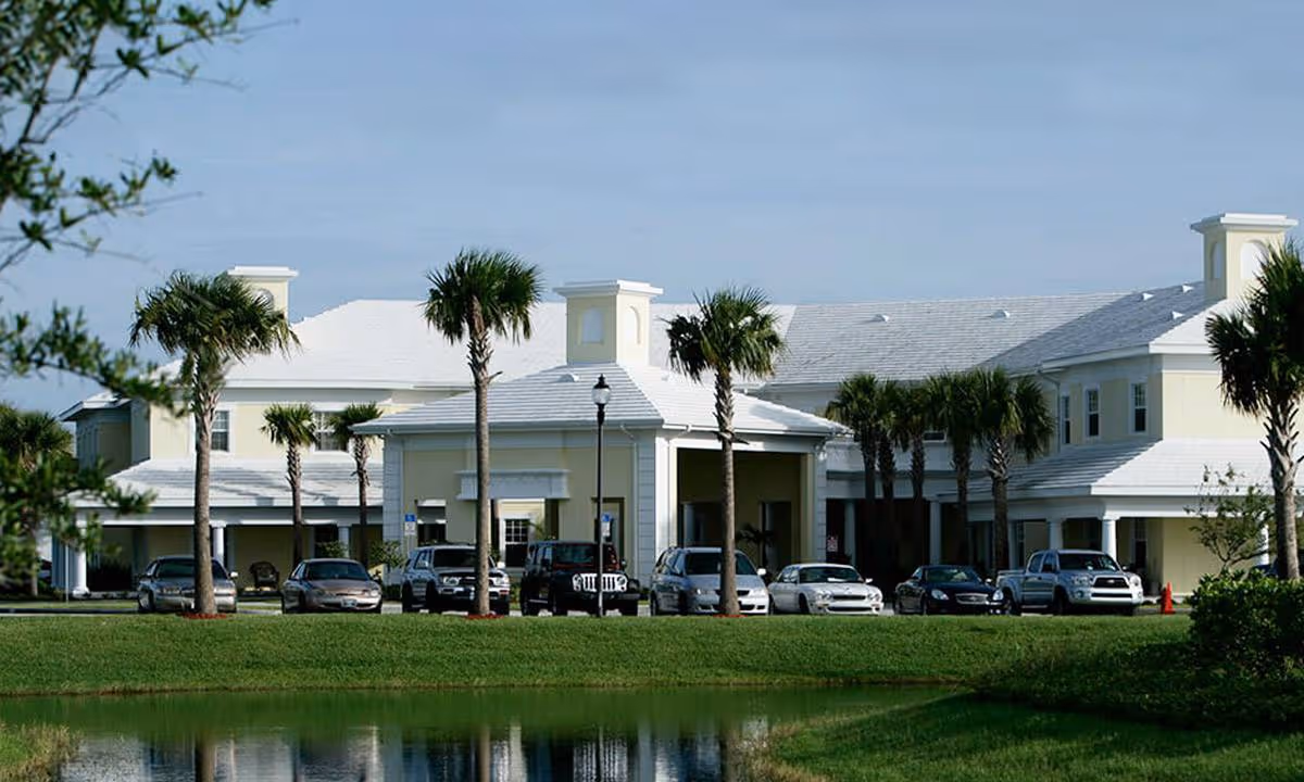 Exterior view of a large, light yellow building with white roofing, surrounded by palm trees and parked cars in front. There is a small body of water reflecting the building and greenery in the foreground under a clear blue sky.