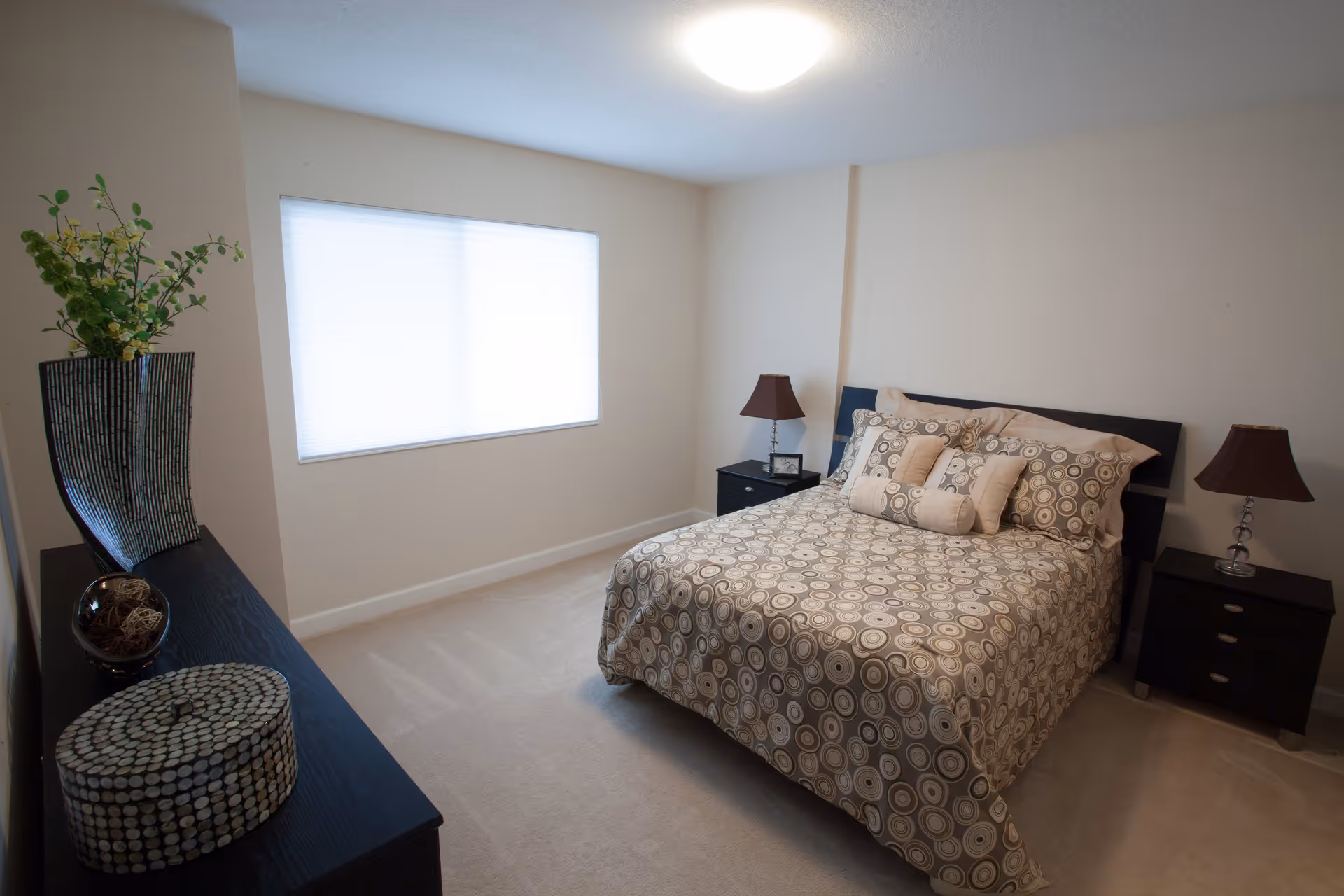 A bedroom with a double bed covered in a patterned bedspread with multiple pillows. There are two dark nightstands on either side of the bed, each with a lamp. A large window with blinds is on the left wall, and a dark dresser with decorative items and a vase with greenery is positioned against the opposite wall.