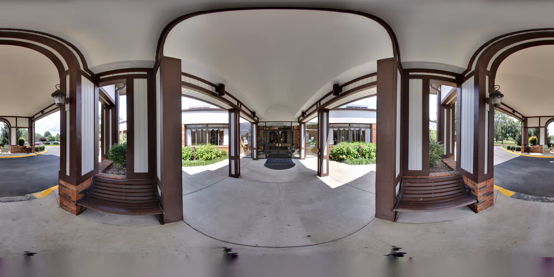 Covered entrance area of The Forum at Brookside senior living facility with brown wooden beams and benches on either side, concrete floor, and glass doors leading inside. Green bushes and trees are visible outside under a partly cloudy sky.