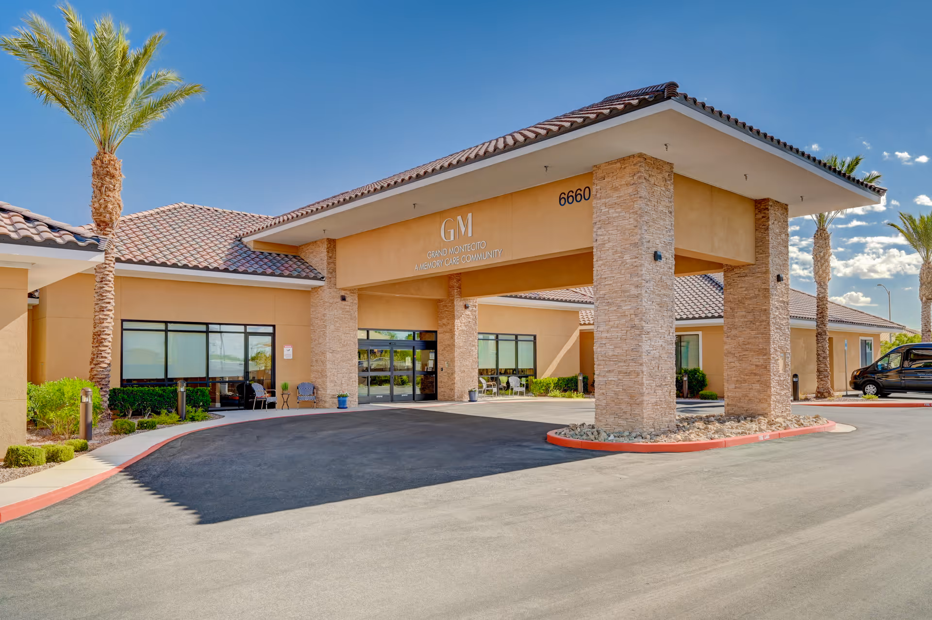 Exterior view of the entrance to Grand Montecito Memory Care community, featuring a covered driveway supported by stone pillars, a tiled roof, palm trees, and clear blue sky.