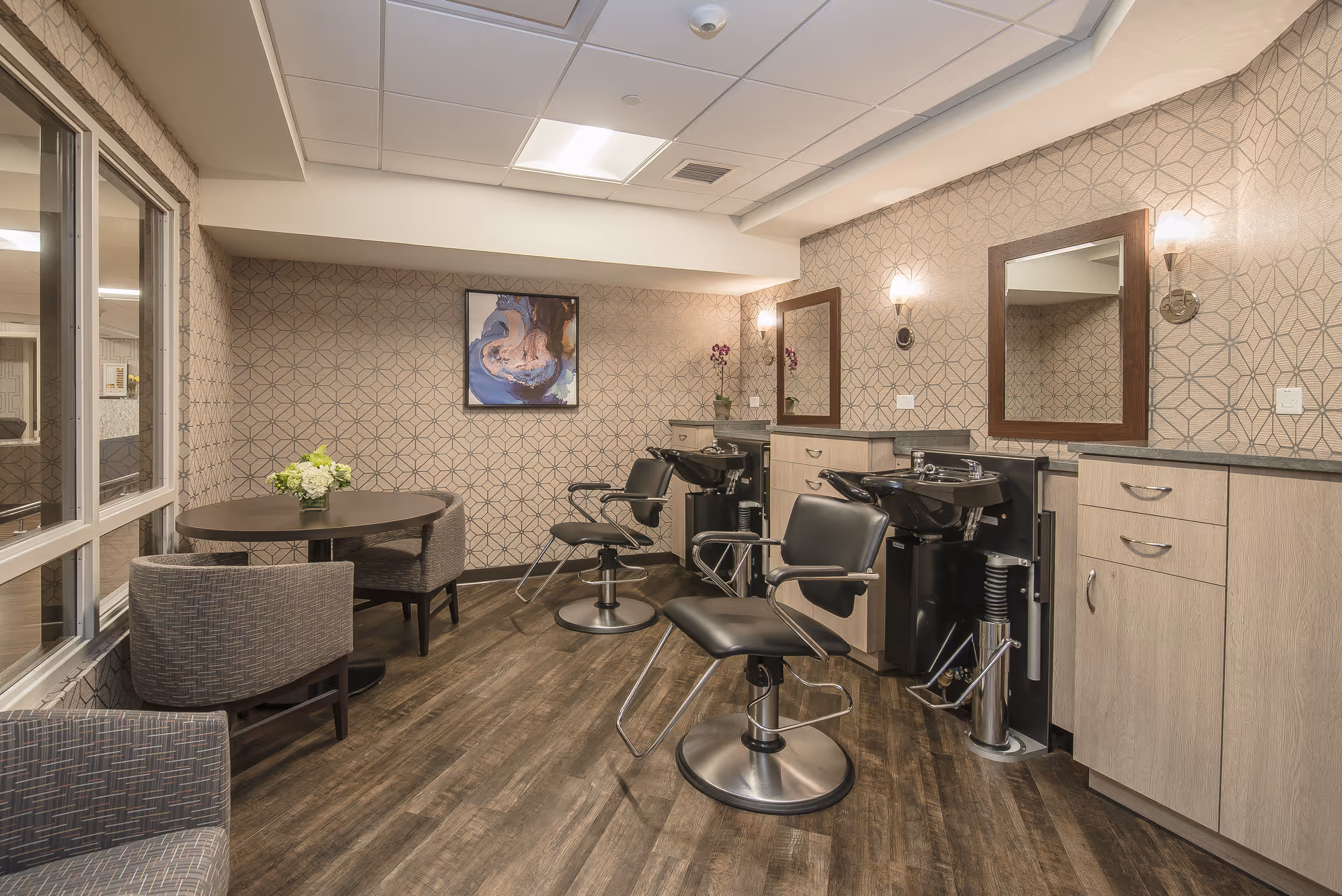 Interior view of a salon area in a senior living facility with two black salon chairs in front of wash basins and mirrors. The room has patterned wallpaper, wooden flooring, a round table with chairs, and a framed abstract painting on the wall.