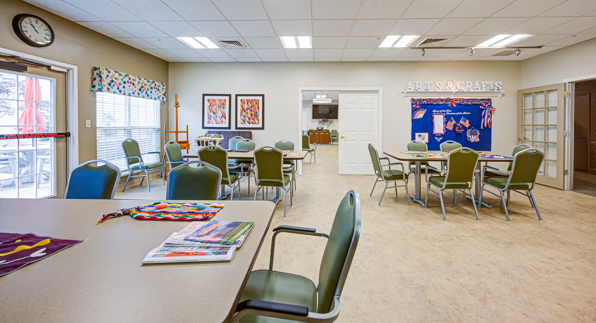 A bright and spacious activity room in a senior living facility with several tables and green chairs arranged for group activities. On the right wall, there is a blue bulletin board labeled 'Arts & Crafts' displaying patriotic-themed decorations. The room has large windows with colorful valances, a clock on the wall, and a door leading to another room with a television and cabinets. Some magazines and a colorful knitted item are on the table in the foreground.