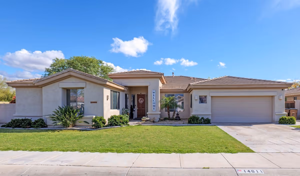 Single-story beige stucco house with a tiled roof, a two-car garage, a front door with a welcome sign, green lawn, and some shrubs under a blue sky with scattered clouds.