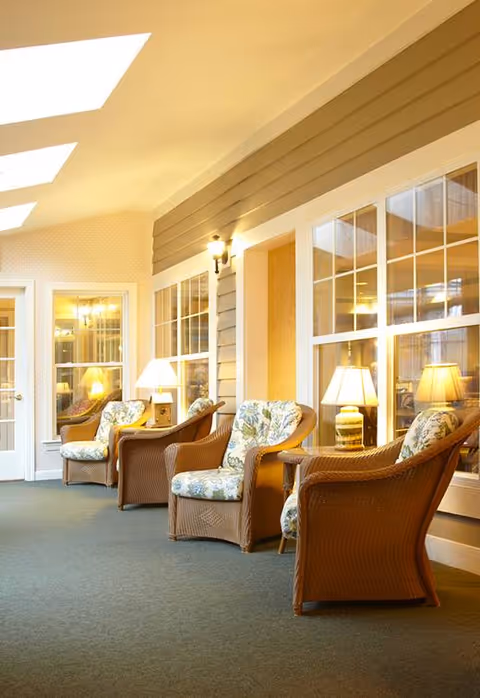 A cozy indoor seating area with wicker armchairs featuring floral cushions, side tables with lamps, large windows, and soft lighting creating a warm atmosphere.