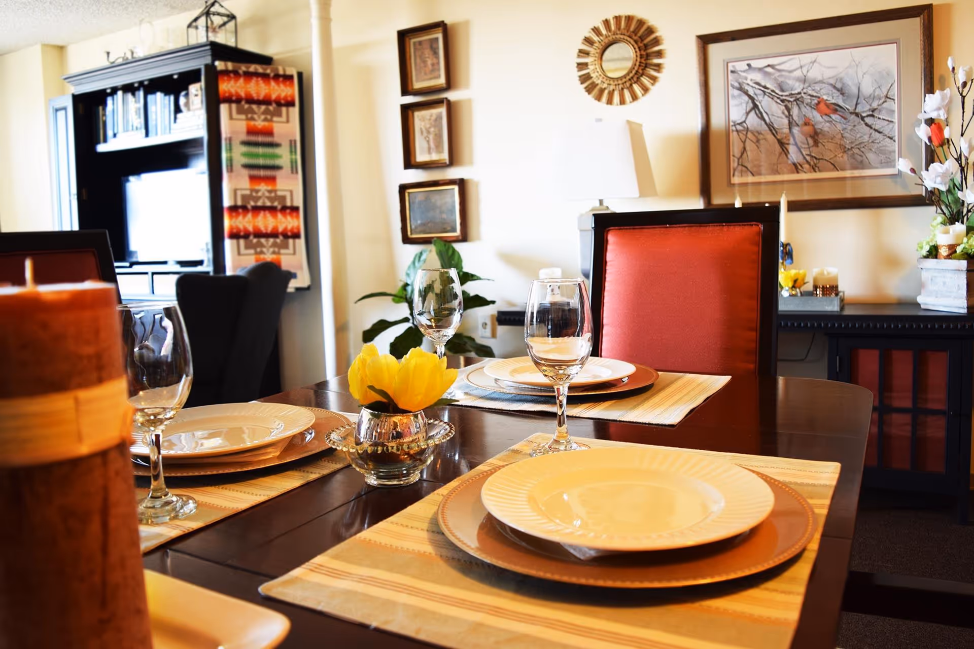 A dining table set for four with plates, wine glasses, and a small vase with a yellow flower in the center. The room has warm lighting, red cushioned chairs, framed artwork on the walls, and a cabinet with decorative items in the background.