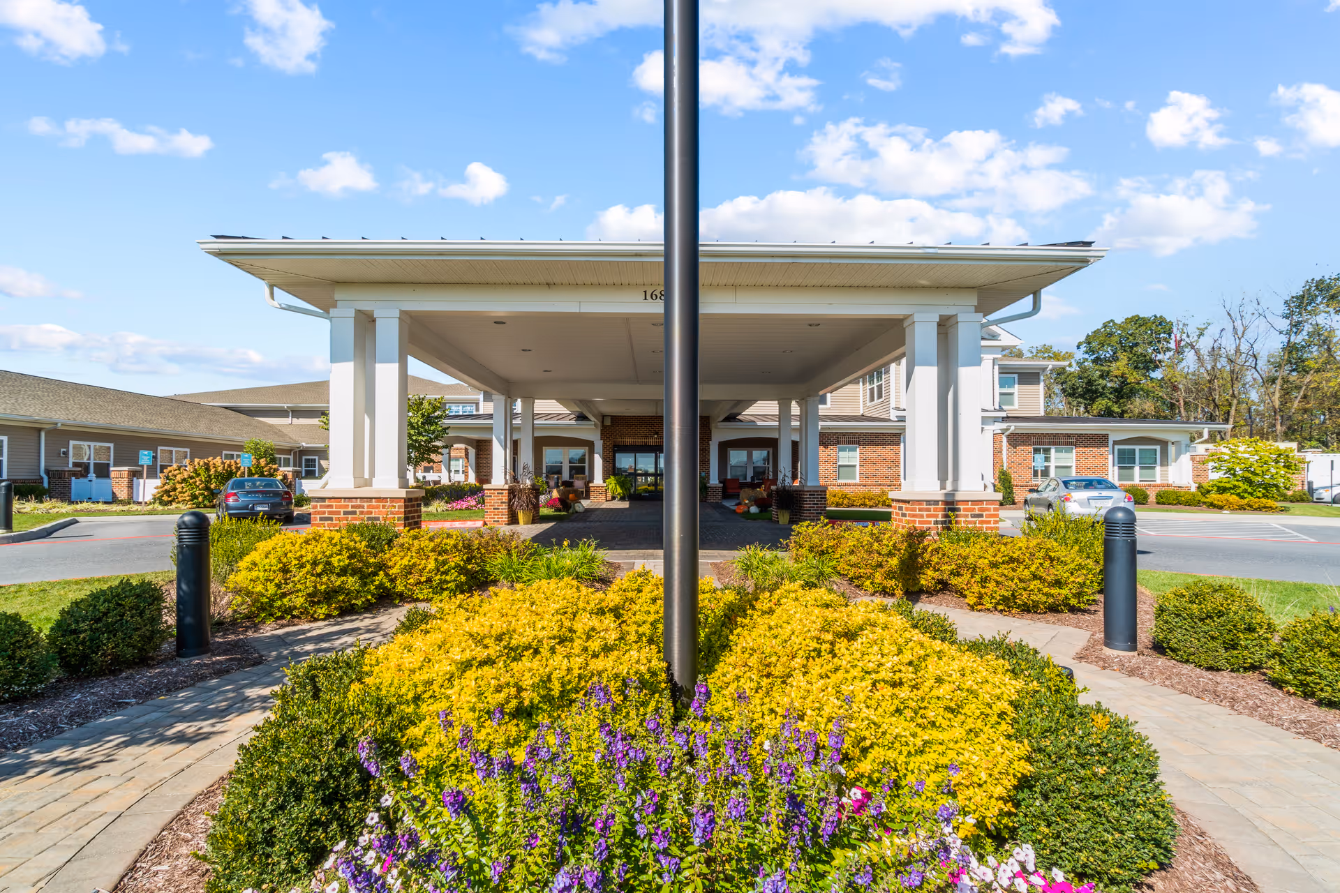 Front entrance of Bellaire at Stone Port Assisted Living & Memory Care facility with a covered drop-off area supported by white columns, surrounded by well-maintained landscaping including yellow and purple flowers, green bushes, and a paved walkway under a blue sky with scattered clouds.