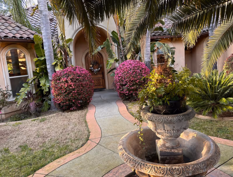 Front entrance of a building with a curved walkway lined with pink flowering bushes and palm trees. There is a large decorative stone planter with green plants in the foreground. The building has arched windows and a wooden door with a wreath.