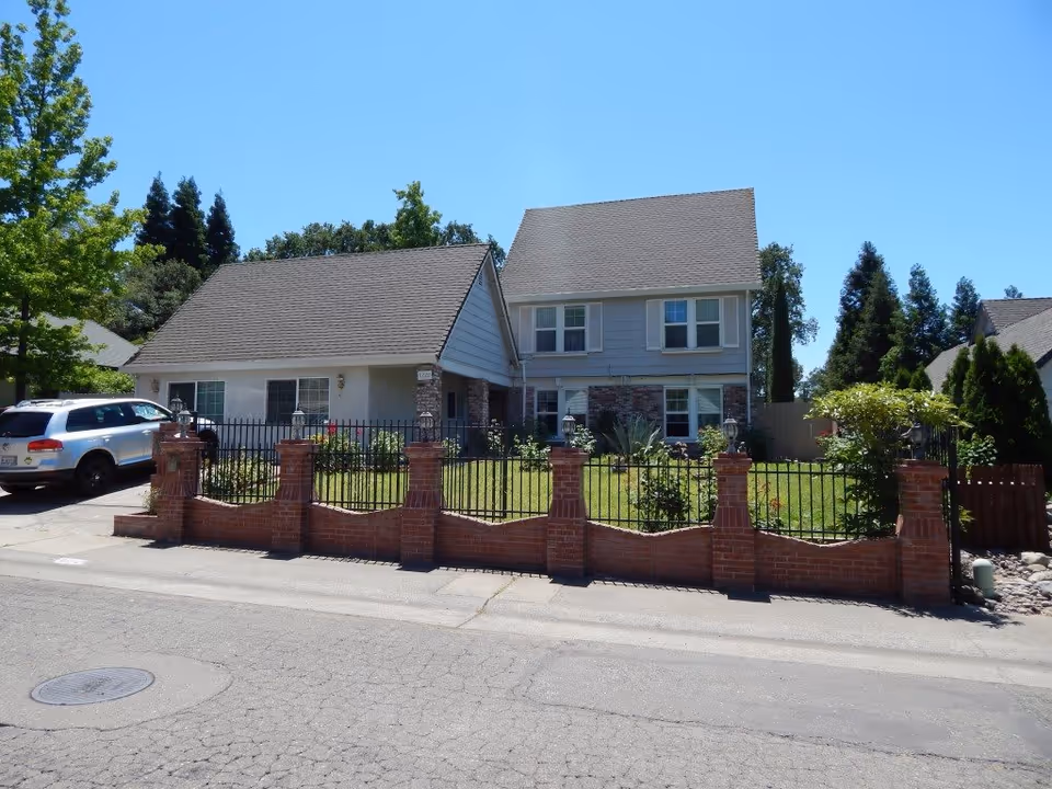 Front exterior view of a two-story residential building with a gray roof and light-colored walls, surrounded by a brick and wrought iron fence. There is a green lawn with some plants and trees, and a white SUV parked in the driveway on the left side. The sky is clear and blue.