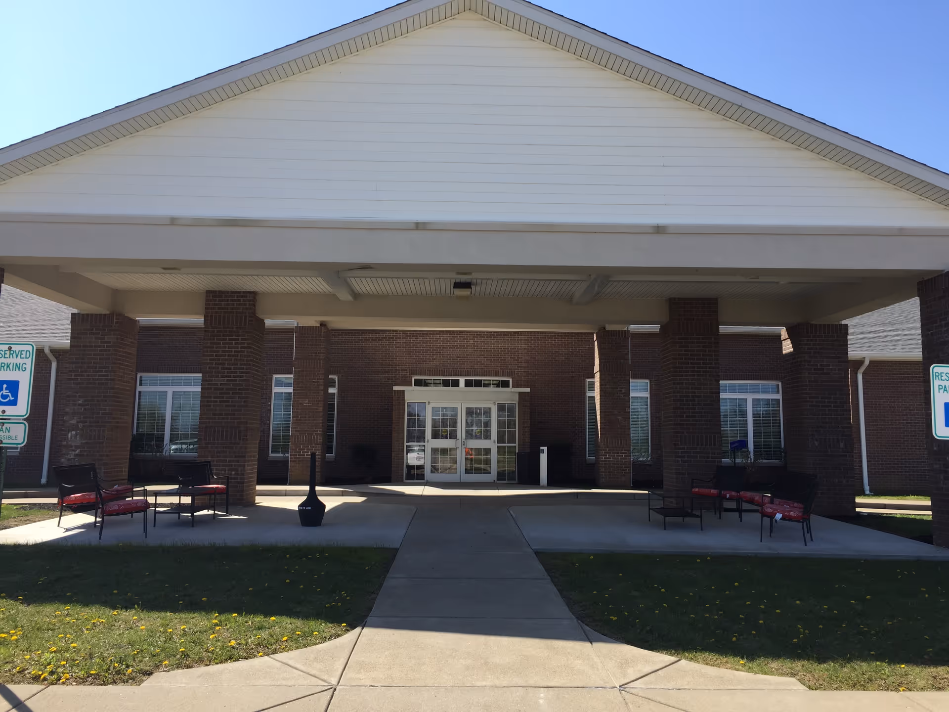 Front entrance of a brick building with a covered driveway supported by brick columns. There are benches with red cushions on either side under the covered area, and reserved parking signs for handicapped parking are visible on both sides. The sky is clear and blue.