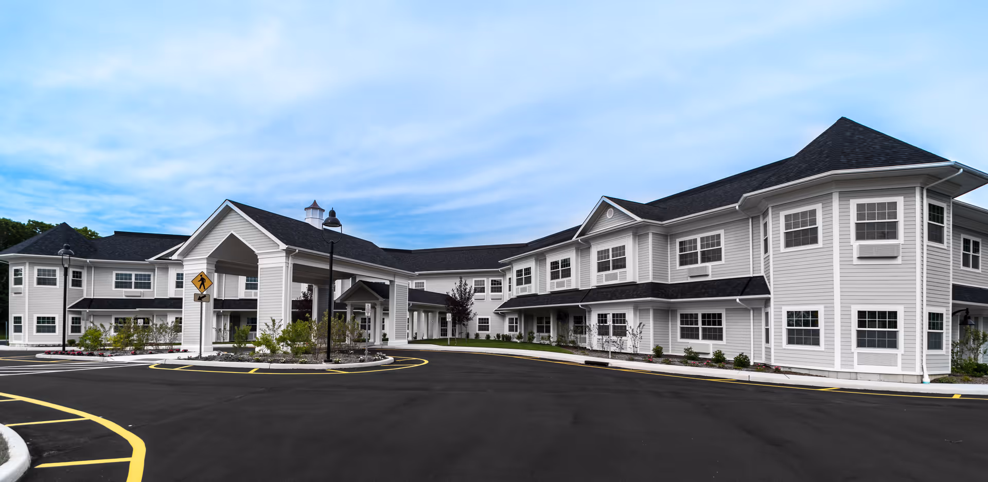 Exterior view of a large, two-story senior living facility building with white siding and black roofing under a partly cloudy sky. The building has multiple windows and a covered entrance with a pedestrian crossing sign nearby. The surrounding area includes a paved driveway and landscaped greenery.
