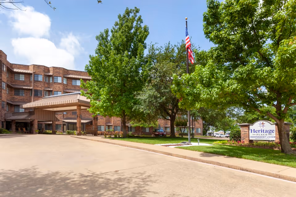 Exterior view of Heritage Place Assisted Living & Memory Care building with a covered entrance driveway, surrounded by green trees and a lawn. An American flag is flying on a flagpole near a sign that reads Heritage Place Independent Living.