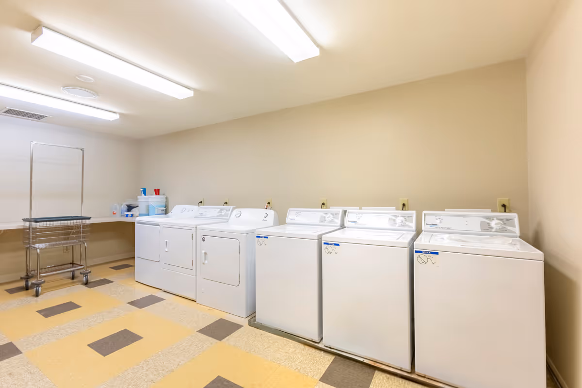 A laundry room with six white washing machines and dryers lined up against a beige wall. There is a metal laundry cart with a handle and a folding table in the corner. The floor has a yellow and brown checkered pattern, and the ceiling has fluorescent lights.