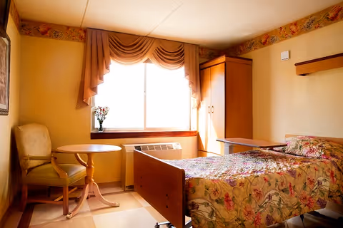 A cozy bedroom in a senior living facility featuring a single bed with floral bedding, a wooden headboard, a small round table with a cushioned chair, a window with draped curtains, a wardrobe, and a vase with flowers on the windowsill.