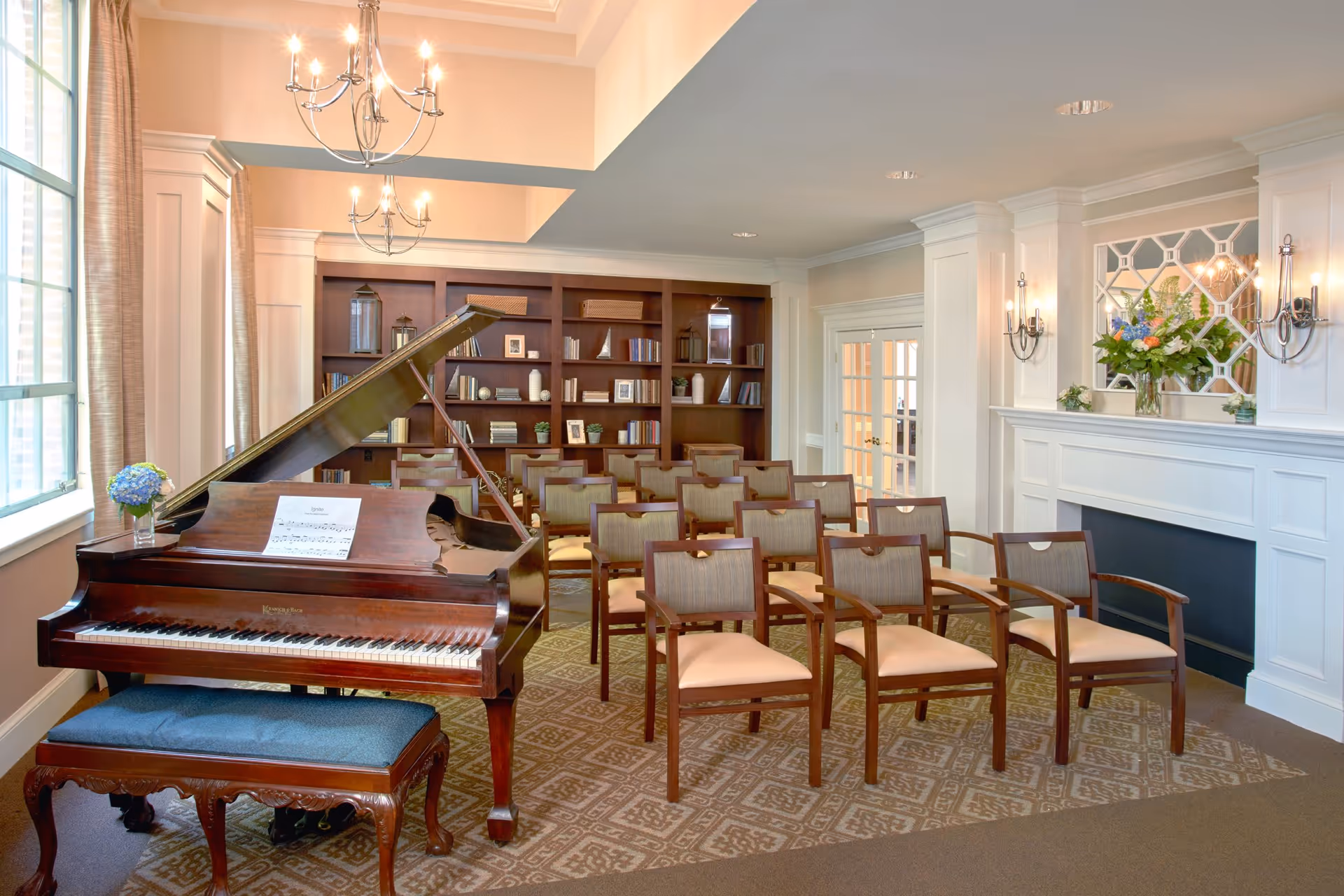A well-lit room with a grand piano and a blue cushioned bench in the foreground. Behind the piano, there are several wooden chairs arranged in rows facing the piano. The room features a large bookshelf filled with books and decorative items, a white fireplace with a mirror above it, and two wall sconces with lit candles. A vase with flowers is placed on the piano and another on the fireplace mantel. The room has large windows with beige curtains and a patterned carpet.